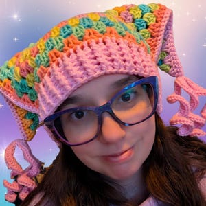 May include: A handmade crochet hat in a granny square pattern. The hat features a pink base with a colorful top section in shades of yellow, green, orange, and pink. The hat has long, curly pink tendrils on each side, adding a whimsical touch.