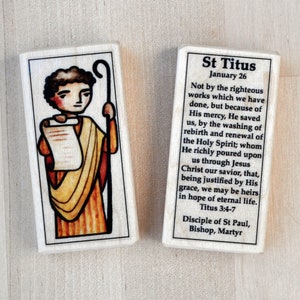 May include: A wooden block with a colorful illustration of Saint Titus holding a scroll and a shepherd's staff. The block also includes a text passage about Saint Titus, including the quote "Not by the righteous works which we have done, but because of His mercy, He saved us."