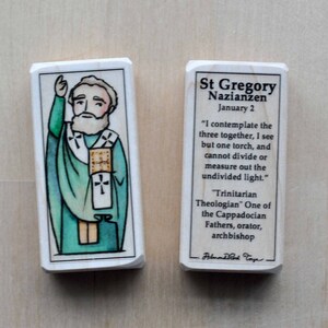 Könnte beinhalten: Zwei rechteckige Holzblöcke. Ein Block zeigt eine handgezeichnete Illustration eines Heiligen in grünen Gewändern, mit weißem Bart und erhobener Hand. Der andere Block hat den Text "St Gregory Nazianzen" und ein Zitat über die Dreifaltigkeit.