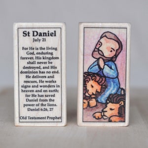 May include: A wooden block with a colorful illustration of Saint Daniel praying with two lions. The block has text that reads "St Daniel, July 21, For He is the living God, enduring forever. His kingdom shall never be destroyed, and His dominion has no end. He delivers and rescues, He works signs and wonders in heaven and on earth; for He has saved Daniel from the power of the lions. Daniel 6:26, 27, Old Testament Prophet."