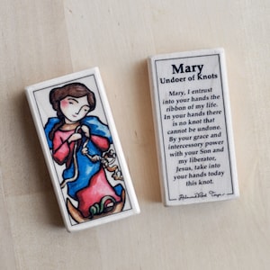 May include: A white rectangular tile with a colorful illustration of Mary, Undoer of Knots. The tile features a prayer to Mary, asking her to undo knots in one's life.