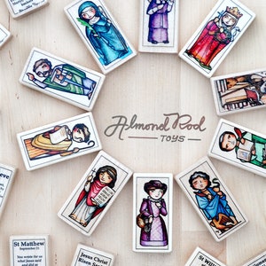May include: A set of colorful wooden dominoes featuring illustrations of saints and religious figures. Each domino has a different saint with a short quote and their name. The dominoes are arranged in a circle on a wooden surface. The text on the dominoes reads "Almond Rod Toys".