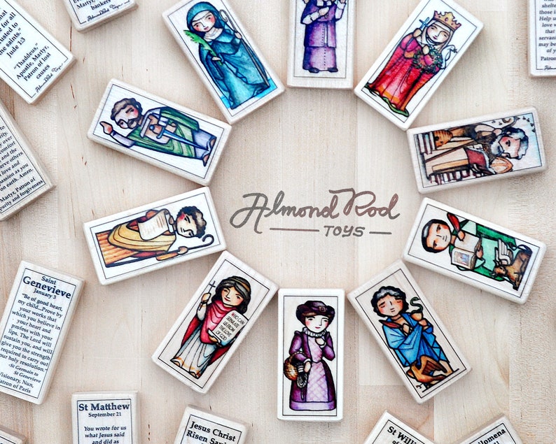 May include: A set of colorful wooden dominoes featuring illustrations of saints and religious figures. Each domino has a different saint with a short quote and their name. The dominoes are arranged in a circle on a wooden surface. The text on the dominoes reads "Almond Rod Toys".