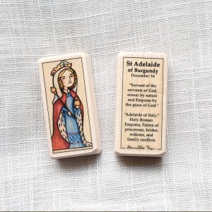 Può includere: Due blocchi rettangolari in legno raffiguranti Santa Adelaide di Borgogna. Un blocco mostra un'illustrazione colorata di un'imperatrice incoronata con un abito blu e un mantello rosso. L'altro blocco riporta un testo nero che descrive la sua vita.