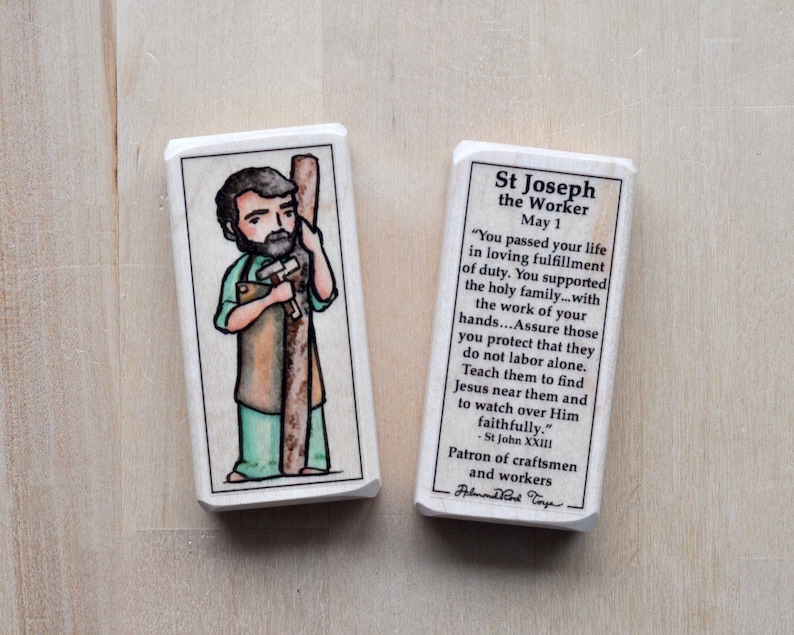 May include: A wooden block with a colorful illustration of Saint Joseph the Worker holding a hammer and a piece of wood. The block has a white background with black text that reads "St Joseph the Worker May 1" and a quote from St John XXIII about the importance of work and supporting one's family. The block is a patron saint of craftsmen and workers.