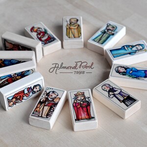 May include: A set of 12 wooden dominoes with colorful illustrations of religious figures. Each domino features a different figure, including a king, a queen, and a priest. The dominoes are arranged in a circle on a light wood surface. The text "Almond Pod Toys" is printed on the surface.