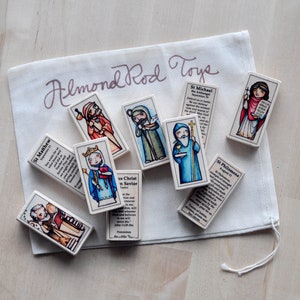 May include: A set of colorful wooden blocks featuring images of saints and religious figures. Each block has a different saint and a short description of their life and work. The blocks are packaged in a white drawstring bag with the text "Almond Rod Toys" printed on it.