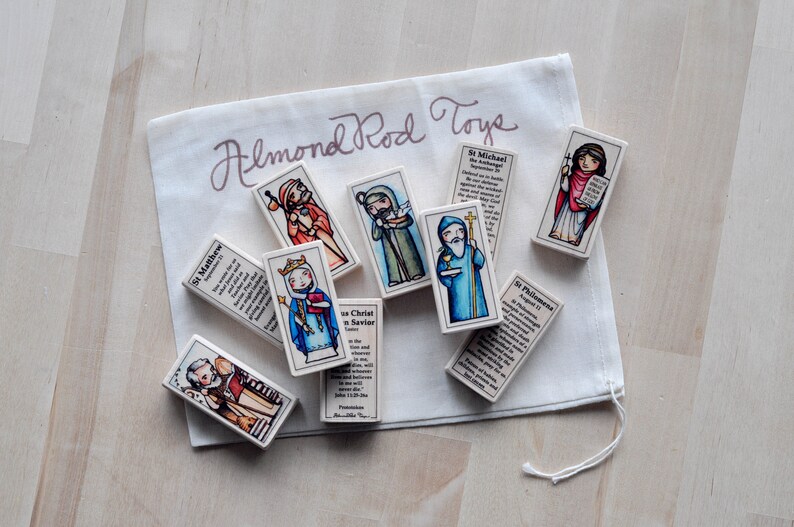 May include: A set of colorful wooden blocks featuring images of saints and religious figures. Each block has a different saint and a short description of their life and work. The blocks are packaged in a white drawstring bag with the text "Almond Rod Toys" printed on it.