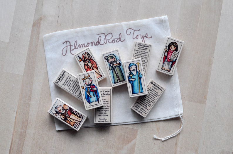 May include: A set of colorful wooden blocks featuring images of saints and religious figures. Each block has a different saint and a short description of their life and work. The blocks are packaged in a white drawstring bag with the text "Almond Rod Toys" printed on it.