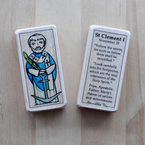 May include: Two wooden Saint Clement I prayer cards. One card features an illustration of Saint Clement holding a palm branch and an anchor. The other card has text that includes quotes and information about Saint Clement.