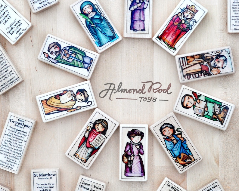 May include: A set of colorful wooden dominoes featuring illustrations of saints and religious figures. Each domino has a different saint with a short quote and their name. The dominoes are arranged in a circle on a wooden surface. The text on the dominoes reads "Almond Rod Toys".