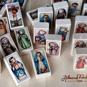 May include: A set of wooden blocks featuring colorful illustrations of religious figures. Each block depicts a different saint or biblical character, including Jesus, Mary, and various angels. The blocks are made of natural wood and have a smooth, polished finish. The blocks are arranged in a random pattern on a wooden surface. The text "Almond Pod Toys" is visible in the bottom right corner of the image.