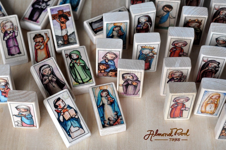 May include: A set of wooden blocks featuring colorful illustrations of religious figures. Each block depicts a different saint or biblical character, including Jesus, Mary, and various angels. The blocks are made of natural wood and have a smooth, polished finish. The blocks are arranged in a random pattern on a wooden surface. The text "Almond Pod Toys" is visible in the bottom right corner of the image.