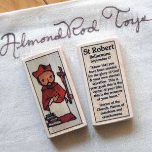 May include: Two wooden saint cards. The left card features a cartoon illustration of Saint Robert Bellarmine, a Catholic priest, holding a book and a cross. The right card has text that reads "St Robert Bellarmine September 17 "Know that you have been created for the glory of God & your own eternal salvation. This is your goal; this is the center of your life; this is the treasure of your heart." Doctor of the Church, Patron of catechists and catechumens"