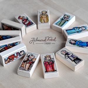 May include: A set of 12 wooden dominoes with colorful illustrations of religious figures. Each domino features a different figure, including a king, a queen, and a priest. The dominoes are arranged in a circle on a light wood surface. The text "Almond Pod Toys" is printed on the surface.