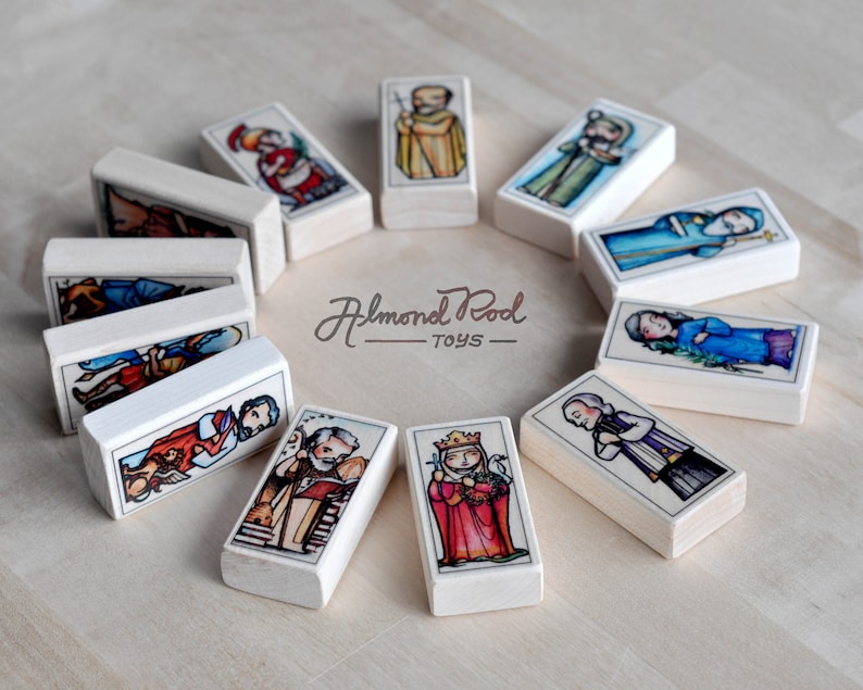 May include: A set of 12 wooden dominoes with colorful illustrations of religious figures. Each domino features a different figure, including a king, a queen, and a priest. The dominoes are arranged in a circle on a light wood surface. The text "Almond Pod Toys" is printed on the surface.