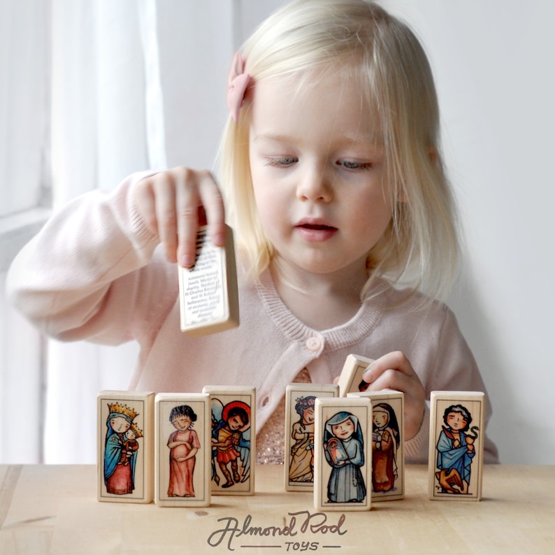May include: A young child plays with a set of colorful wooden blocks featuring images of saints. The blocks are arranged in a row on a wooden table. The blocks are part of a set of religious toys for children.