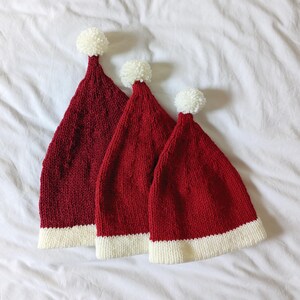 May include: Three red knit Santa hats with white trim and white pom-poms. The hats are arranged from largest to smallest, with a textured knit pattern. The hats are on a white background.