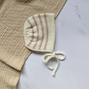May include: A white knitted baby bonnet with pink stripes and a tie. The bonnet is lying on a beige fabric.