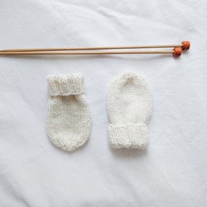 May include: Two white knitted baby mittens with wooden knitting needles.
