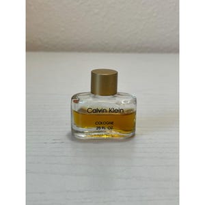 May include: A small, clear glass cologne bottle with a gold cap. The bottle contains a golden-colored liquid and features the text "Calvin Klein" and "COLOGNE .25 FL OZ". The bottle is rectangular with rounded edges.
