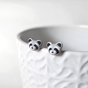 May include: A pair of small, raccoon-shaped stud earrings. Each earring features a gray and white face with black accents, including the eyes and ears. The earrings are set against a white background, highlighting their design.