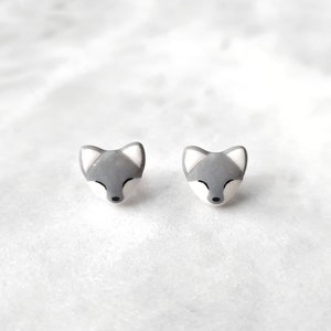 Puede incluir: Un par de pendientes de botón con forma de zorro gris y blanco. Los pendientes presentan una cara de zorro detallada con los ojos cerrados y una pequeña nariz negra. Las orejas del zorro son blancas y la cara es gris.