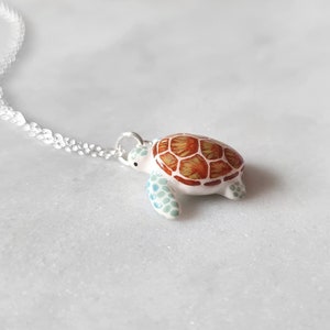 May include: A silver chain necklace with a white ceramic turtle pendant. The turtle has a brown and orange shell with green accents.