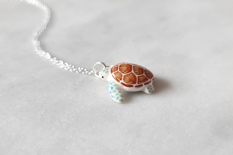 May include: A silver chain necklace with a white ceramic turtle pendant. The turtle has a brown and orange shell with green accents.