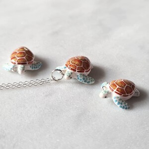 May include: A silver chain necklace with a small, white turtle pendant. The turtle has a brown shell with blue accents. There are two additional smaller turtles, one on each side of the pendant.