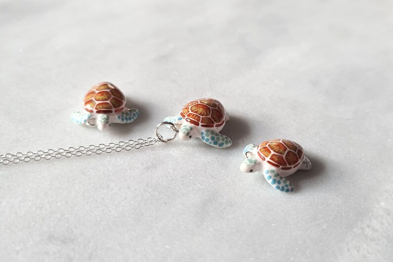 May include: A silver chain necklace with a small, white turtle pendant. The turtle has a brown shell with blue accents. There are two additional smaller turtles, one on each side of the pendant.