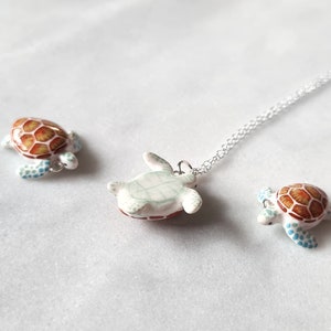 May include: A silver chain necklace with a ceramic turtle pendant. The turtle is painted white with brown and green accents. Two smaller ceramic turtles are also pictured, one with a brown and white shell and the other with a brown and blue shell.
