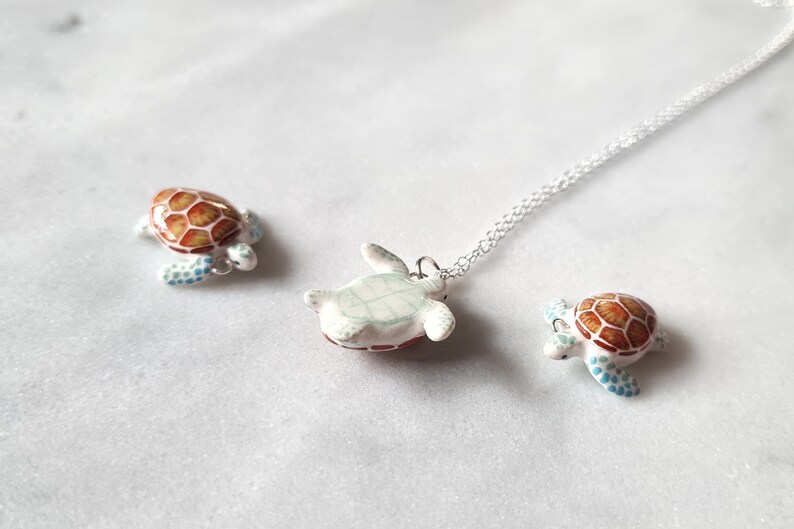 May include: A silver chain necklace with a ceramic turtle pendant. The turtle is painted white with brown and green accents. Two smaller ceramic turtles are also pictured, one with a brown and white shell and the other with a brown and blue shell.