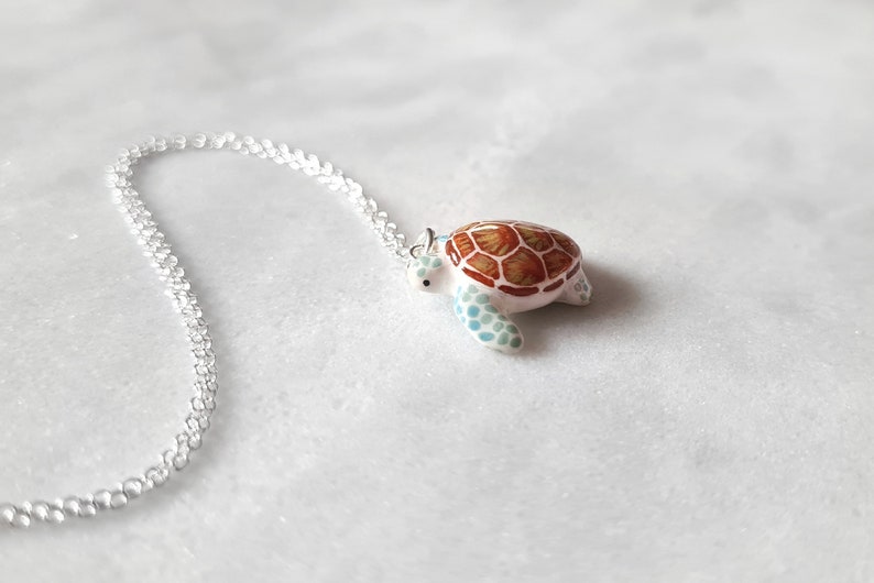 May include: A silver chain necklace with a white and brown turtle pendant. The turtle has a detailed shell with brown and orange markings and blue accents.