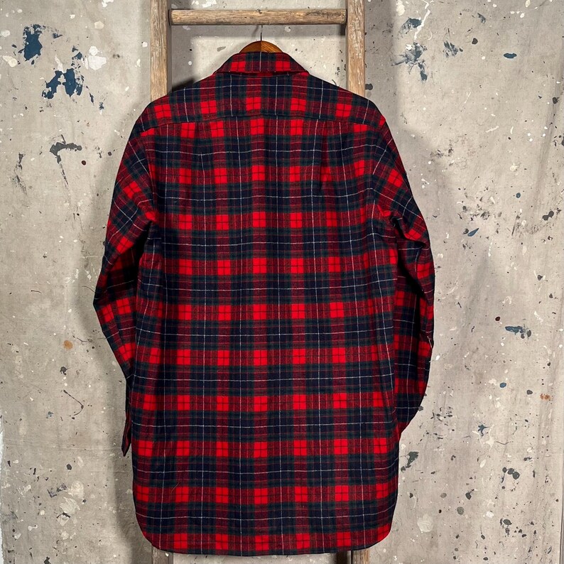 Puede incluir: Una camisa de franela a cuadros rojos y negros cuelga de una percha de madera. La camisa est&aacute; hecha de una tela suave y tejida y presenta un dise&ntilde;o cl&aacute;sico con botones. El patr&oacute;n de cuadros es un dise&ntilde;o tradicional.