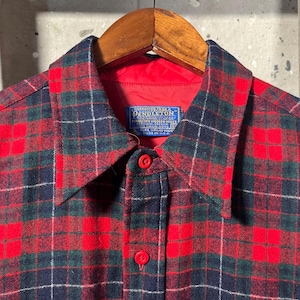 Puede incluir: Una camisa a cuadros roja, verde y azul marino con botones. La camisa tiene un forro rojo y un cuello. La etiqueta dice "Pendleton Woolen Mills". La camisa est&aacute; colgada en una percha de madera.