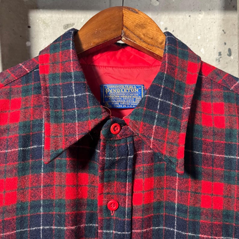 Puede incluir: Una camisa a cuadros roja, verde y azul marino con botones. La camisa tiene un forro rojo y un cuello. La etiqueta dice "Pendleton Woolen Mills". La camisa est&aacute; colgada en una percha de madera.