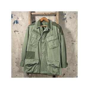 May include: Olive green military-style jacket with multiple front pockets and patches. The jacket is hanging on a wooden hanger against a weathered background. The jacket has a collared neckline and button-down closure.