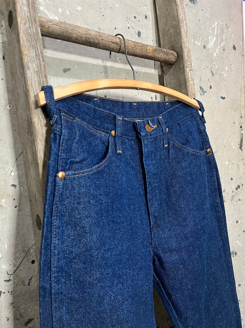 May include: A pair of dark blue denim jeans hanging on a wooden hanger. The jeans have a classic five-pocket style and a straight leg.
