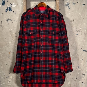 Puede incluir: Una camisa de franela a cuadros rojos y negros con mangas largas, dos bolsillos en el pecho y una botonadura. La camisa est&aacute; colgada de una percha de madera sobre un fondo neutro.