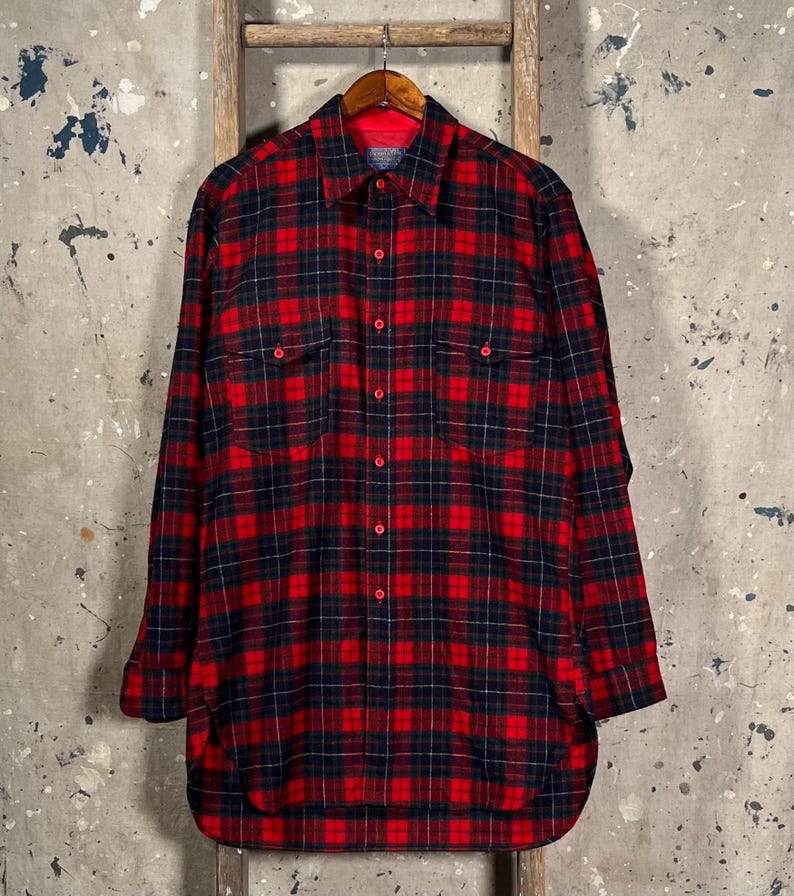 Puede incluir: Una camisa de franela a cuadros rojos y negros con mangas largas, dos bolsillos en el pecho y una botonadura. La camisa est&aacute; colgada de una percha de madera sobre un fondo neutro.