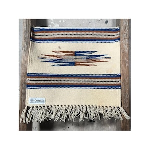 May include: Cream-colored woven textile with horizontal stripes in brown and blue. A central geometric design in brown and blue is featured. The textile has a fringe along the bottom edge and a label that reads "Ortega's".