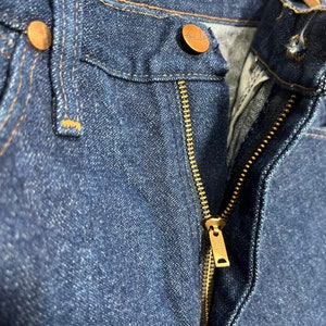 May include: Close-up of a pair of blue denim jeans with a brass zipper and a copper button. The jeans have a classic five-pocket style.