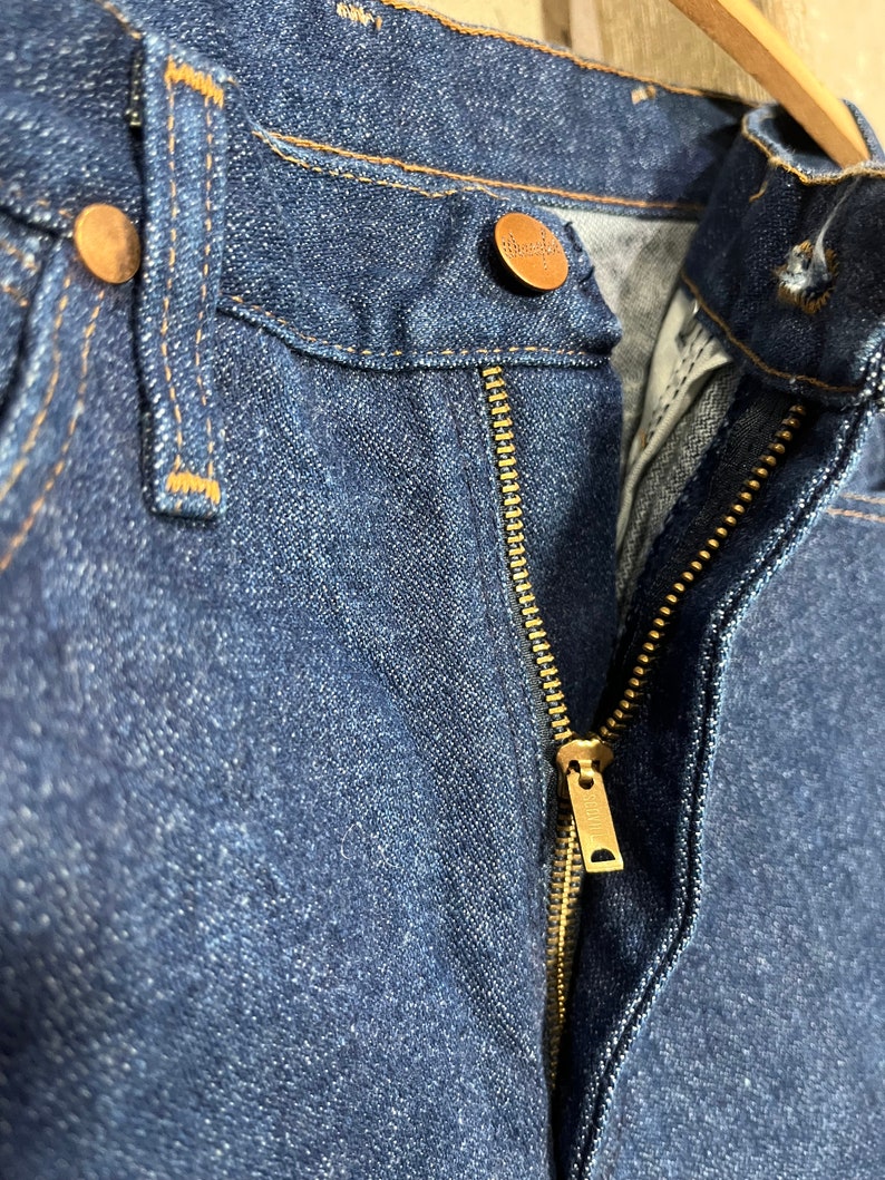 May include: Close-up of a pair of blue denim jeans with a brass zipper and a copper button. The jeans have a classic five-pocket style.