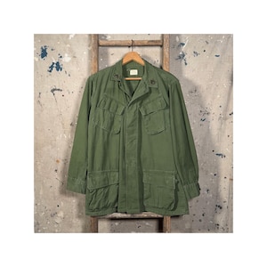 Può includere: Giacca verde oliva in stile militare con più tasche frontali e colletto. La giacca è appesa a una scala di legno su uno sfondo grigio e bianco screziato. La giacca ha una chiusura con bottoni e maniche lunghe.