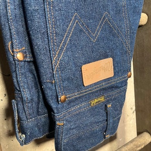 Puede incluir: Un par de pantalones vaqueros de mezclilla azul oscuro con un parche de cuero marr&oacute;n que dice "Wrangler" en el bolsillo trasero. Los vaqueros tienen un estilo cl&aacute;sico de cinco bolsillos y una pierna recta.