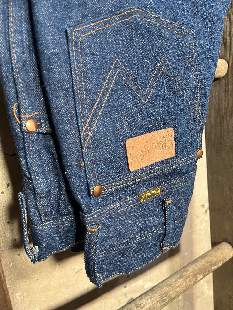 Puede incluir: Un par de pantalones vaqueros de mezclilla azul oscuro con un parche de cuero marr&oacute;n que dice "Wrangler" en el bolsillo trasero. Los vaqueros tienen un estilo cl&aacute;sico de cinco bolsillos y una pierna recta.