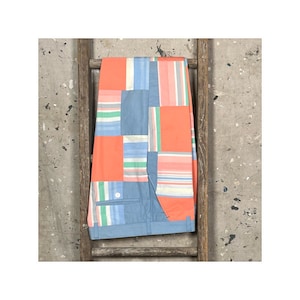 Può includere: Pantaloncini patchwork nei toni del blu, corallo e motivi a righe. I pantaloncini sono piegati e mostrati su una scala di legno su uno sfondo grigio screziato. I pantaloncini hanno una chiusura con bottoni e tasche.