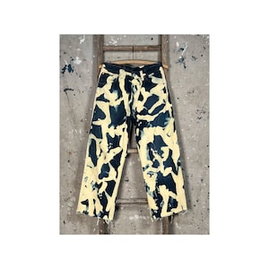 Puede incluir: Vaqueros de mezclilla azul oscuro con un patrón abstracto blanqueado en crema. Los vaqueros tienen un diseño clásico de cinco bolsillos y un dobladillo sin rematar deshilachado. Cuelgan de una escalera de madera contra una pared texturizada.