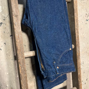 Puede incluir: Un par de pantalones vaqueros de mezclilla azul oscuro con cierre de cremallera y trabilla de cintur&oacute;n de cuero marr&oacute;n. Los vaqueros est&aacute;n colgados de una escalera de madera.